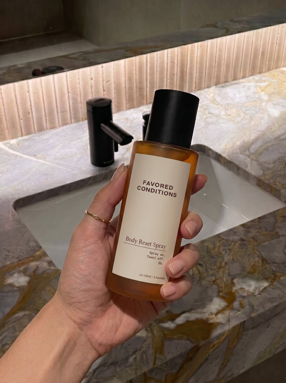 A hand holding a Favored Conditions Body Reset Spray bottle over a marble sink in a warmly lit bathroom